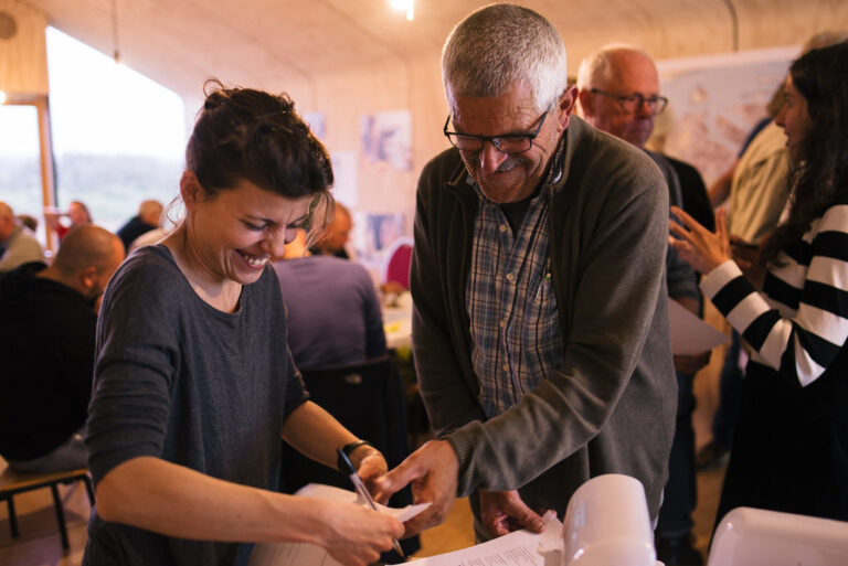 Een foto van een vrouw en een man tijdens een bijeenkomst. De vrouw lacht en pakt een boekje aan van de man die rechts van haar staat.